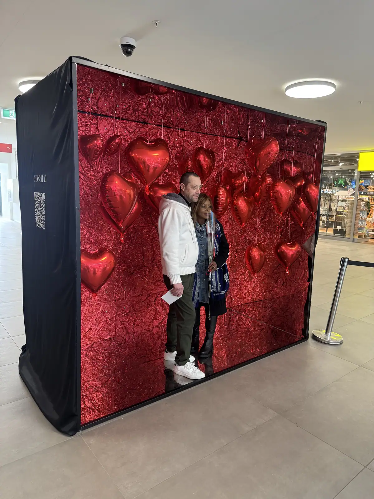 Un couple pose dans la Cosam-Box aux cœurs rouges, le photobooth installé devant, en galerie marchande